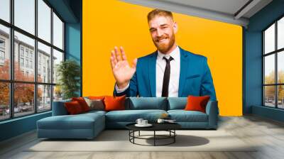 young red head businessman smiling happily and cheerfully, waving hand, welcoming and greeting you, or saying goodbye against orange background Wall mural