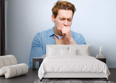 young red head man feeling ill with a sore throat and flu symptoms, coughing with mouth covered against blue wall Wall mural