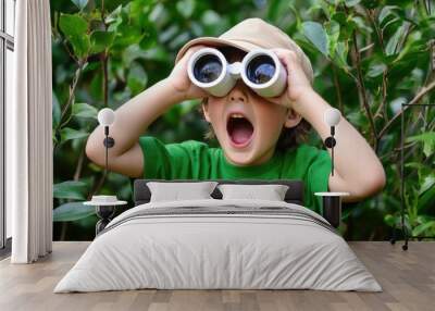 A young boy wearing a hat and green shirt, looking through binoculars amidst lush greenery, shows excitement. Wall mural