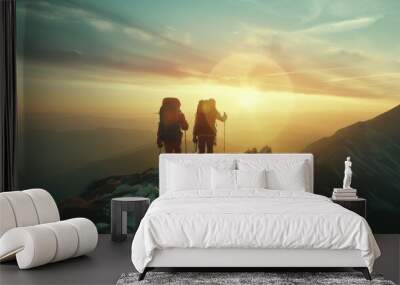 Two hikers are standing on a mountaintop watching the sunset. The sky is orange and the sun is setting behind the mountains. Wall mural