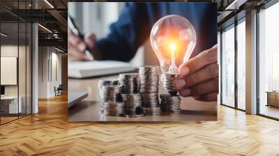 Businessman holding and putting lightbulb on coins stack on table for saving energy and saving money concept. Wall mural