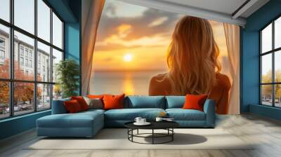 Close up and back view of an attractive woman standing in front of windows at a tropical resort hotel room, with a lovely view of ocean, sunsetting at the blurred background. Wall mural