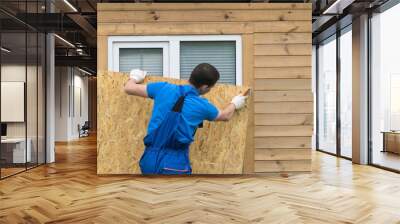 a man obstructs a window with a large piece of plywood before a natural disaster, a hurricane Wall mural