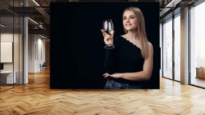 Charming dreamy blonde in casual clothing standing over black background with glass of white wine. Young woman enjoying beverage in studio. Wall mural