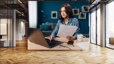 Confident young lady with curly hair working on laptop and reading paper documents at home. Cheerful woman in denim shirt sitting at table and using computer. Wall mural