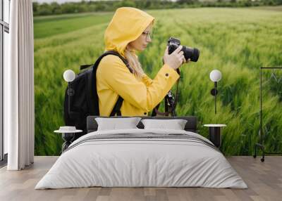 Side view of female photographer using tripod and professional camera for shooting green wheat field. Charming woman in jacket with hood and eyeglasses standing at countryside with black backpack. Wall mural