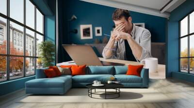 Young man in casual outfit rubbing his eyes because of long working day with portable computer. Bearded freelancer feeling tired while using laptop at home. Wall mural