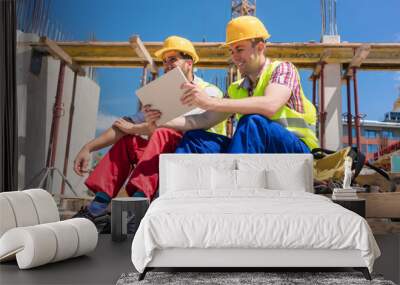 Side view of two workers, reading online information or watching a video on a tablet PC during break at work on the construction site of a contemporary building Wall mural