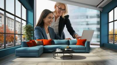 Two happy busy female employees working together using computer planning project. Middle aged professional business woman consulting teaching young employee looking at laptop sitting at desk in office Wall mural