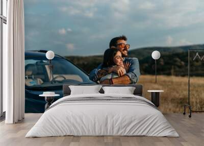 A happy young man with a hand on his girlfriend's shoulder looking away while standing by a car against a clear sky during sunset. Wall mural