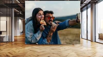 Loving couple taking a selfie outdoors, leaning on the hood of their car in the evening, eating fast food Wall mural