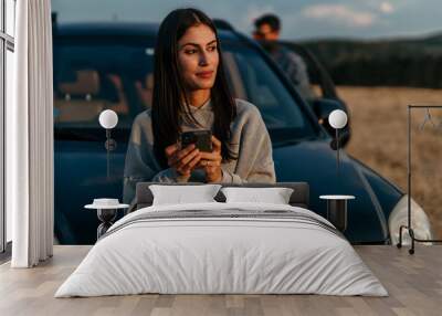 Young cheerful woman using a smartphone next to her car on the hills Wall mural