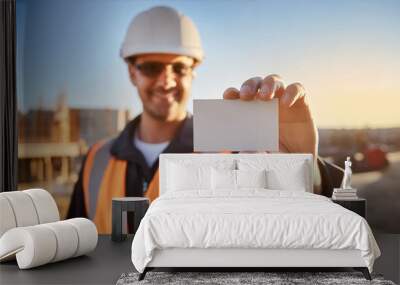 Worker man with helmet, safety vest at construction site, holding an empty business card close to the camera, focus on the business card mockup. Wall mural