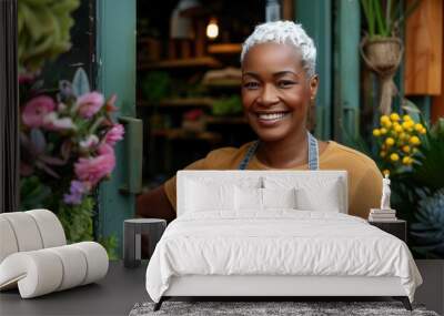 Black woman with a flower shop apron is smiling and posing for a picture. The shop is filled with various plants and flowers, including a large bouquet of pink flowers. Small business. Wall mural