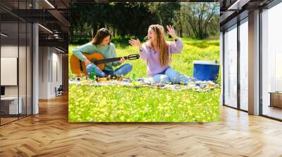 Two women playing a guitar, singing and dancing, doing a picnic sitting on a blanket in a yellow flower meadow. Wall mural