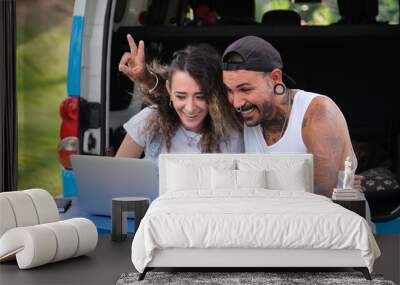 Young tattooed couple greet and talk in a laptop video call during a van trip. Wall mural