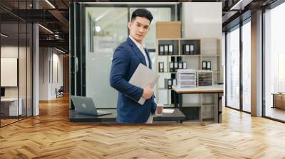 Confident Asian man with a smile standing holding notepad and tablet at the modern office. Wall mural