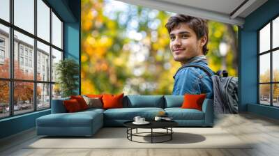  A male university student with a backpack, smiling and enjoying a sunny day in a park. The student is dressed casually and appears relaxed amidst the greenery and natural surroundings. Wall mural