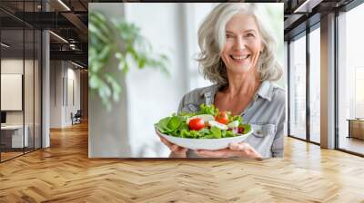 A happy elderly woman holding a bowl of fresh organic green vegetable salad in a light kitchen. Eating healthy food. Concept of Diet, Fitness, Healthy lifestyle, vegetarianism.  Wall mural