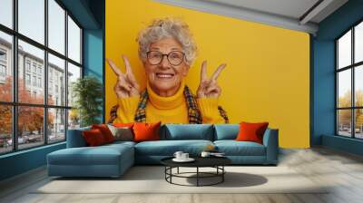 Happy senior woman in glasses, wearing yellow, making peace sign with both hands, bright background

 Wall mural