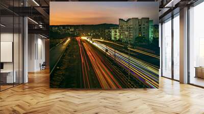 Slow shutter speed image of light trails, beautiful sunset sky , buildings and flowing traffic on motorway. Wall mural