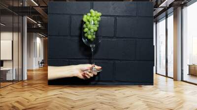 Close-up of female hand holding a wine glass with green grapes inside on background of black brick wall. Wall mural