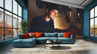 Night portrait of young cheerful guy working on laptop in dark room with garlands on table at home. Wall mural