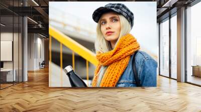 Portrait of young blonde teenager girl, wearing blue denim jacket, with orange scarf and plaid cap, holding a black steel thermo eco water bottle. Wall mural