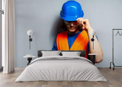 Studio portrait of young construction worker wearing safety equipment; blue hard hat, transparent goggles and orange vest on the background of grey wall. Wall mural