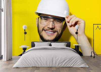 Studio portrait of young smiling man architect, builder engineer, wearing black sweater, touching his white construction safety helmet and glasses, isolated on yellow background. Wall mural