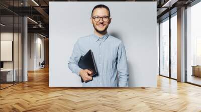 Studio portrait of young smiling man with laptop in hand, on white background. Wearing blue shirt and eyeglasses. Wall mural