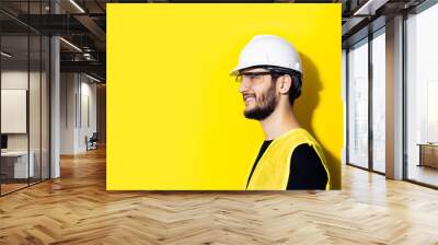 Studio profile portrait of young smiling man architect, builder engineer, wearing white construction safety helmet, glasses and yellow jacket isolated on yellow background. Wall mural