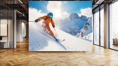 A skier in orange clothing speeds down a snowy mountain slope, leaving a trail of powder behind them. The sun shines brightly in the blue sky, with fluffy clouds and snow-capped mountains Wall mural