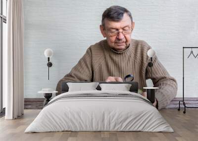 Elderly man reads composition of food product with magnifier. Fine print on can. Hands of old senior with magnifying glass on wooden table, close up, copy space Wall mural