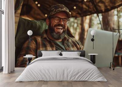 A bearded man in casual attire laughs heartily while using a laptop in an outdoor setting, reflecting joy, relaxation, and digital connectivity. Wall mural