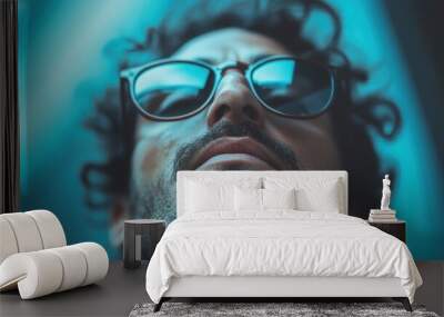 A bearded man sporting sunglasses confidently poses against a blue background, showcasing modern style, confidence, and a sense of assuredness and poise. Wall mural