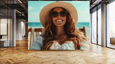 A joyful woman smiles while sitting on a beach wearing stylish sunglasses and a straw hat, basking in the sunny daylight by the tranquil sea. Wall mural