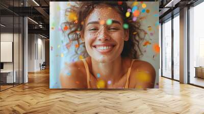 A joyful young woman is smiling with colorful confetti on her face and in the air around her Wall mural