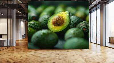 A single halved avocado with a glossy seed stands out among whole avocados, amidst a natural and fresh environment Wall mural