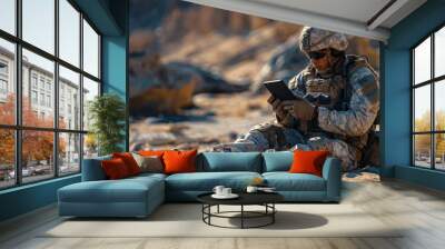 A soldier, clad in military camouflage and armed with a rifle, sits on the ground in an outdoor setting, gazing intently at a tablet, surrounded by the tools of war as he awaits orders from his comma Wall mural