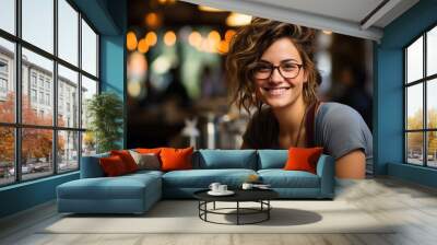 A stylish woman with a warm smile, adorned in glasses, stands confidently in an indoor restaurant, radiating joy and charm as she captures the camera's attention Wall mural