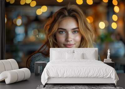 A young woman with striking blue eyes and lovely curls smiles at a cozy cafe, laptop in front of her Wall mural