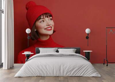 Portrait of a joyful woman wearing a red knit hat with a pom-pom and matching sweater, looking to the side against a red background, capturing a warm and stylish winter vibe.
 Wall mural