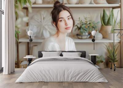 A woman is sitting in a white robe in front of a shelf full of plants. She is smiling and looking at the camera Wall mural