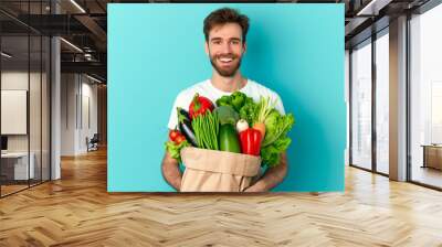 A man is smiling and holding a bag full of vegetables. The vegetables include broccoli, carrots, and peppers Wall mural