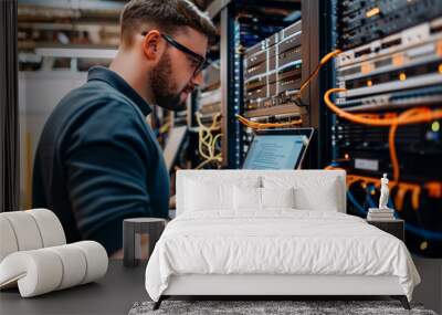 A man is working on a laptop in front of a large network of cables. He is focused on his work, possibly troubleshooting a problem or configuring a system Wall mural