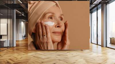 Senior woman is applying makeup to her face. She is smiling and looking at the camera. Concept of self-care and confidence Wall mural