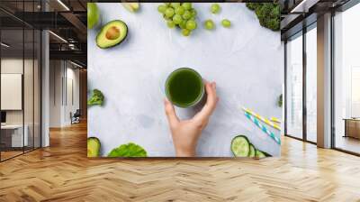 Frame flatlay with various green fruits and vegetables: lettuce, cucumber, avocado, broccoli, grapes, apples etc and woman's hand holding green juice on cement background. Healthy lifestyle concept Wall mural