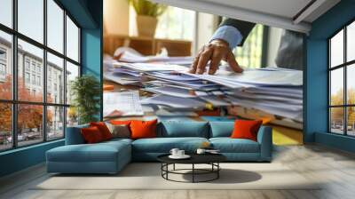 An office desk cluttered with document reports, with a businessman's hand reaching for one Wall mural