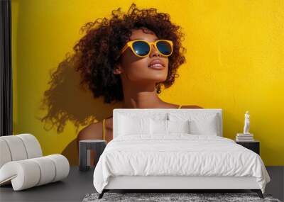 Positive young black woman is standing in front of yellow wall, wearing sunglasses and smiling Wall mural